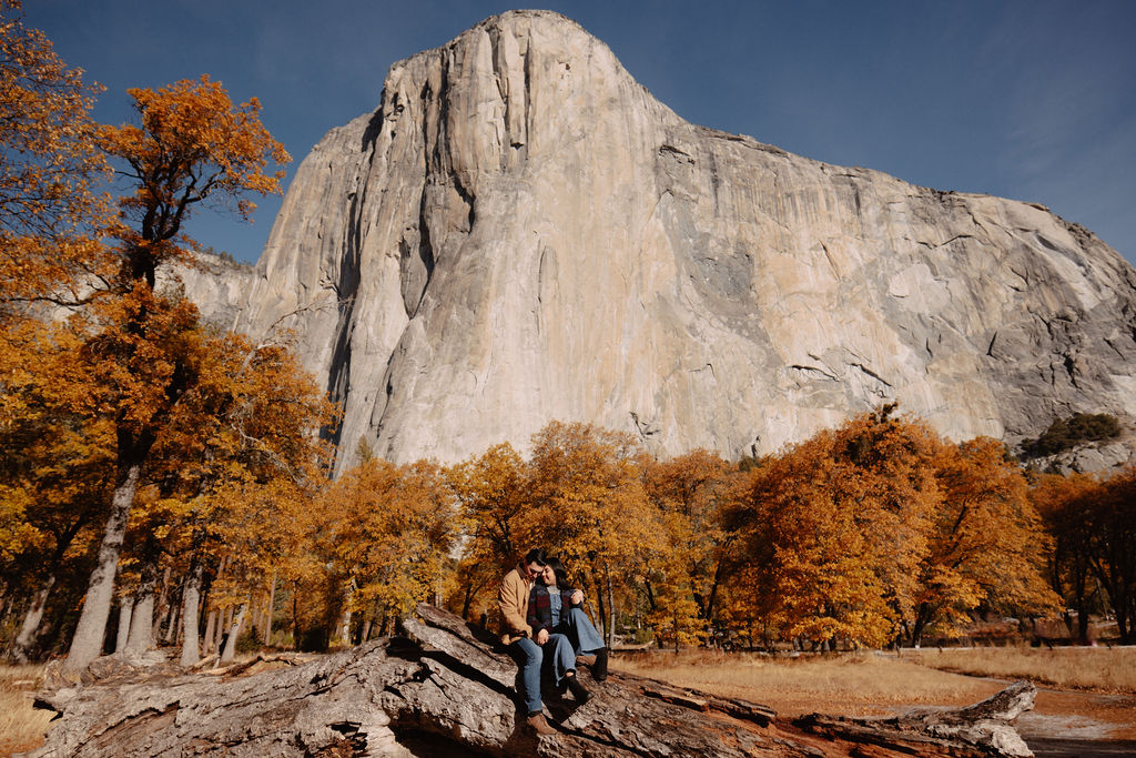 A couple sits on a fallen log in front of a large rock formation, surrounded by autumn trees for their yosemite fall photos