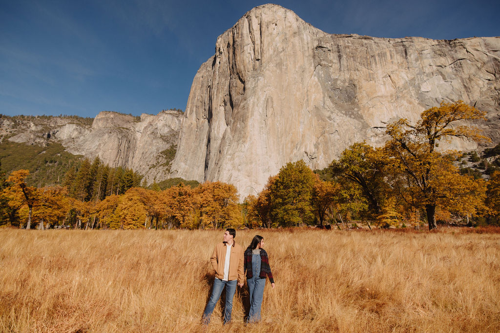 A couple sits on a fallen log in front of a large rock formation, surrounded by autumn trees for their yosemite fall photos