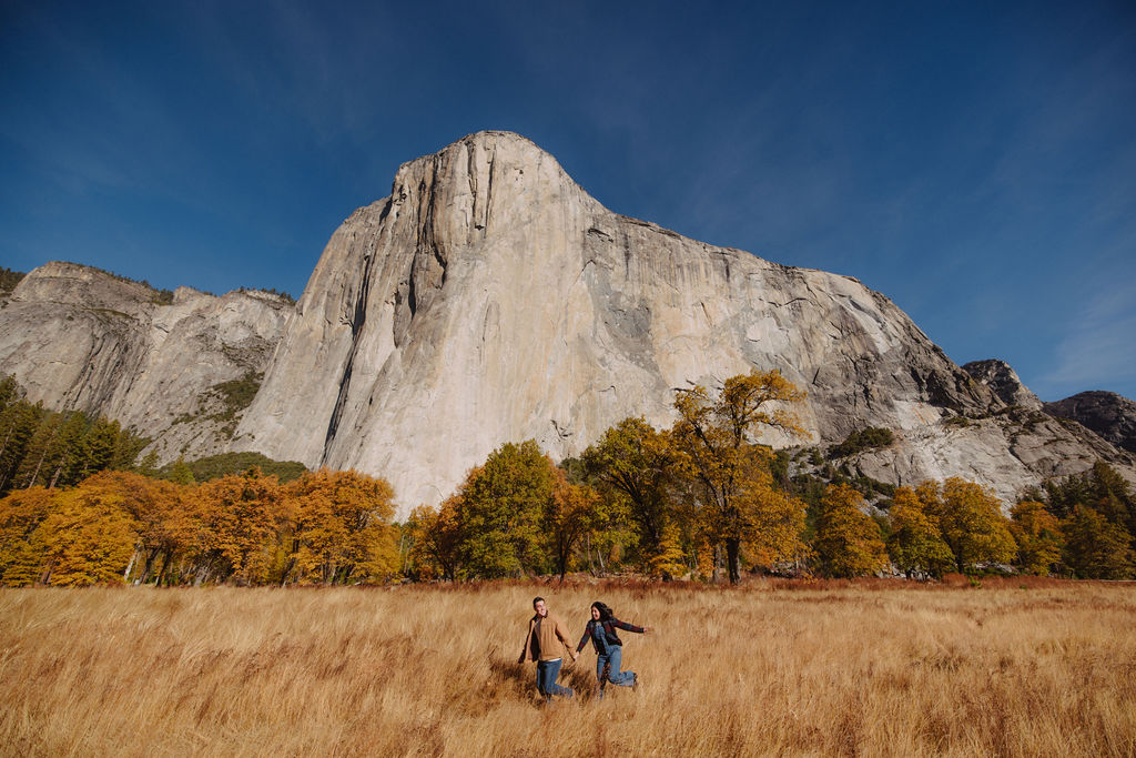 A couple sits on a fallen log in front of a large rock formation, surrounded by autumn trees for their yosemite fall photos