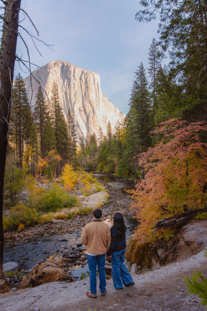 Two people stand facing a scenic view of El Capitan in Yosemite, surrounded by a forest with autumn foliage and a flowing river for their yosemite fall photos