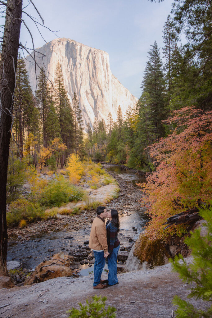 A couple kisses by a reflective lake, surrounded by trees with autumn colors and mountains in the background.