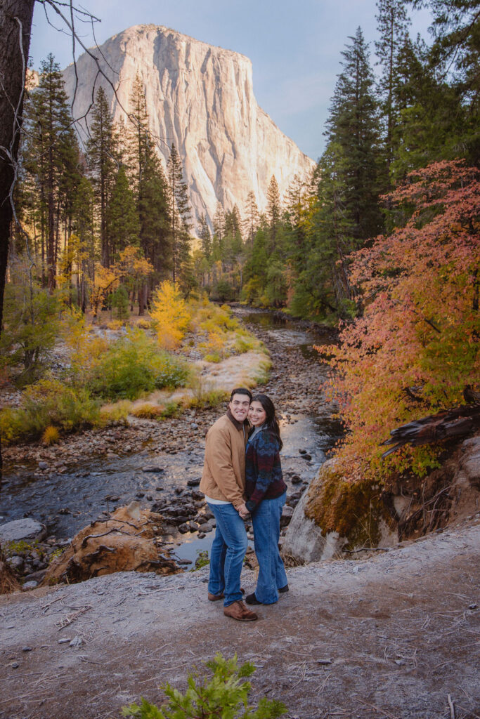 A couple kisses by a reflective lake, surrounded by trees with autumn colors and mountains in the background.