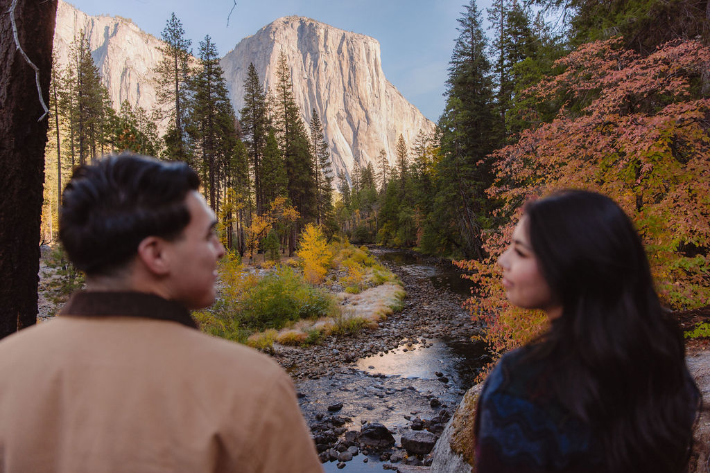 Two people stand near a river, looking at a large cliff surrounded by trees with autumn foliage for their Yosemite fall photos