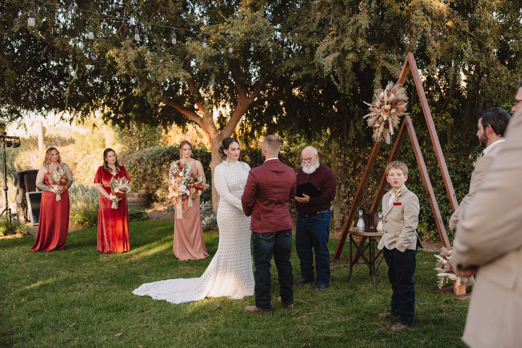 A couple is exchanging vows outdoors under a tree. The bride is in a white dress, and the groom is in a maroon jacket. A person holding a bouquet stands nearby. The Difference Between an Elopement and a Traditional Wedding