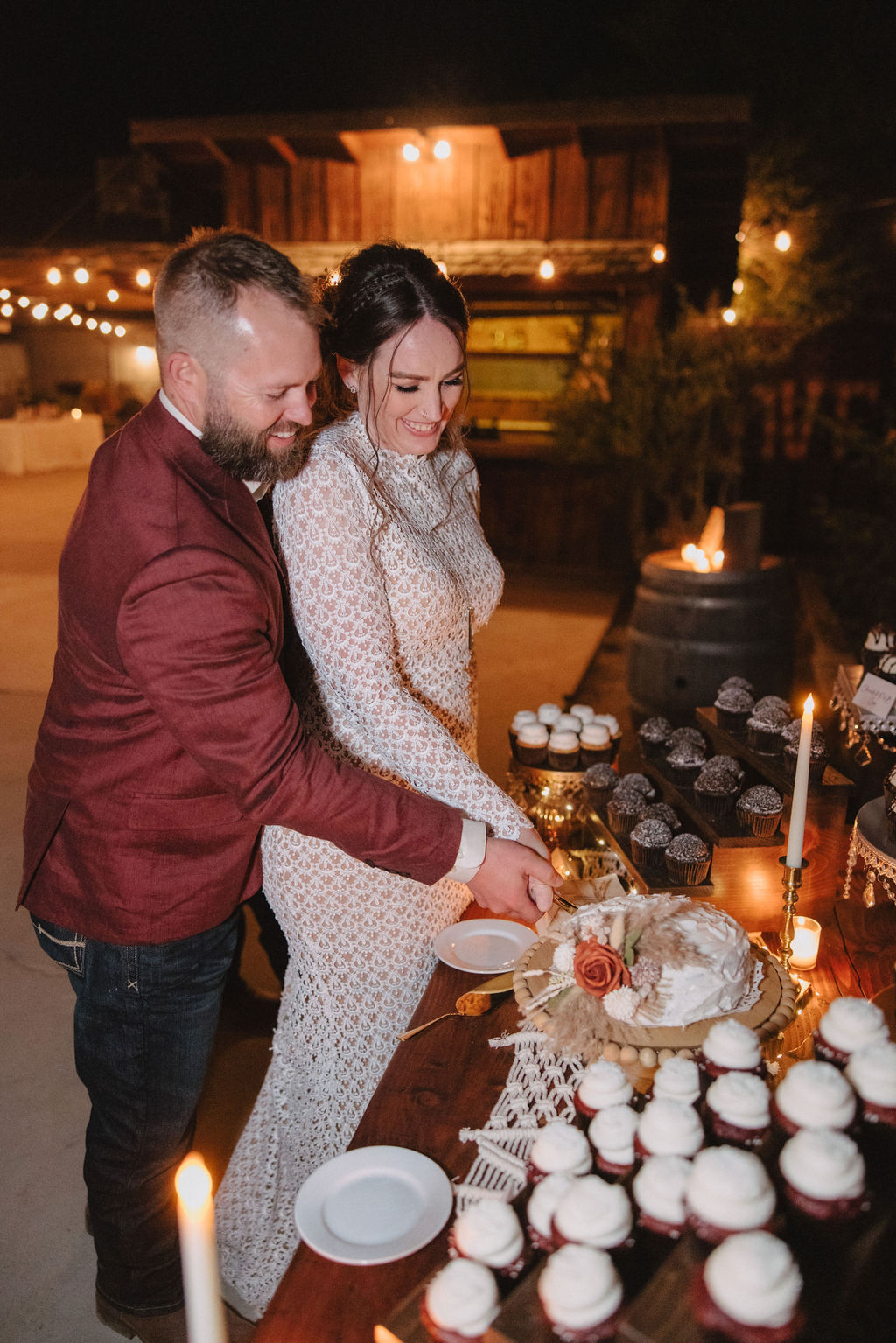 A couple is cutting a cake together at an outdoor event. The table is adorned with cupcakes and lit candles.