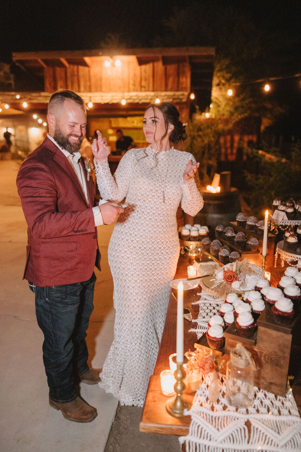 A couple is cutting a cake together at an outdoor event. The table is adorned with cupcakes and lit candles.