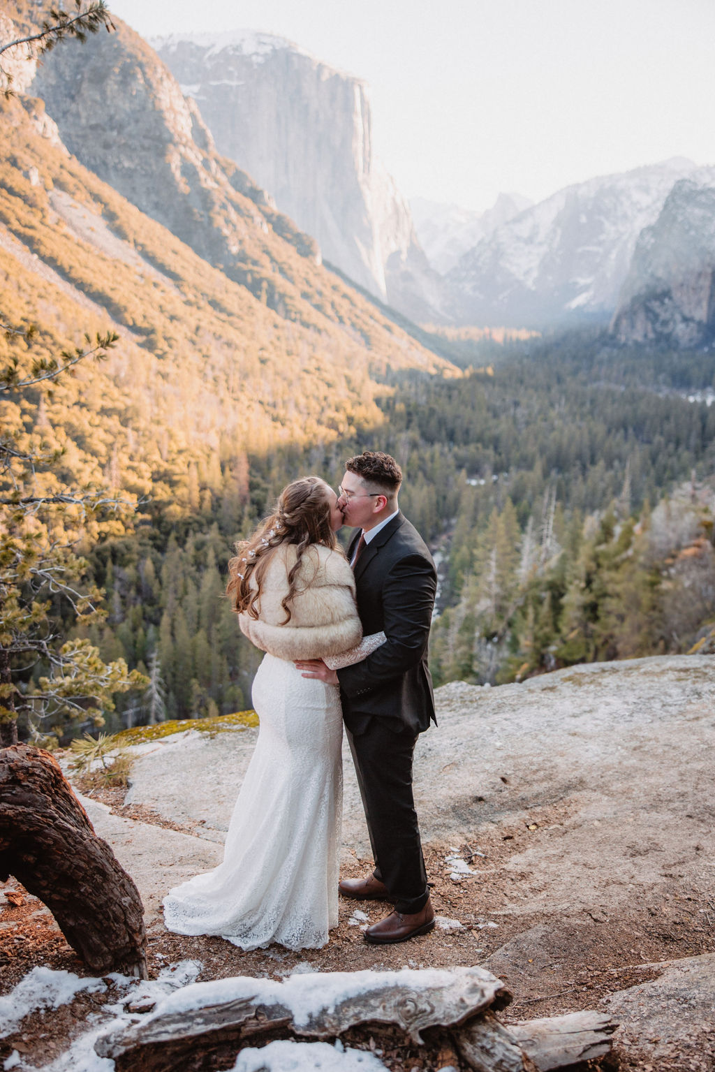 A couple embraces on a rocky overlook with a forest and mountain landscape in the background for their yosemite winter wedding