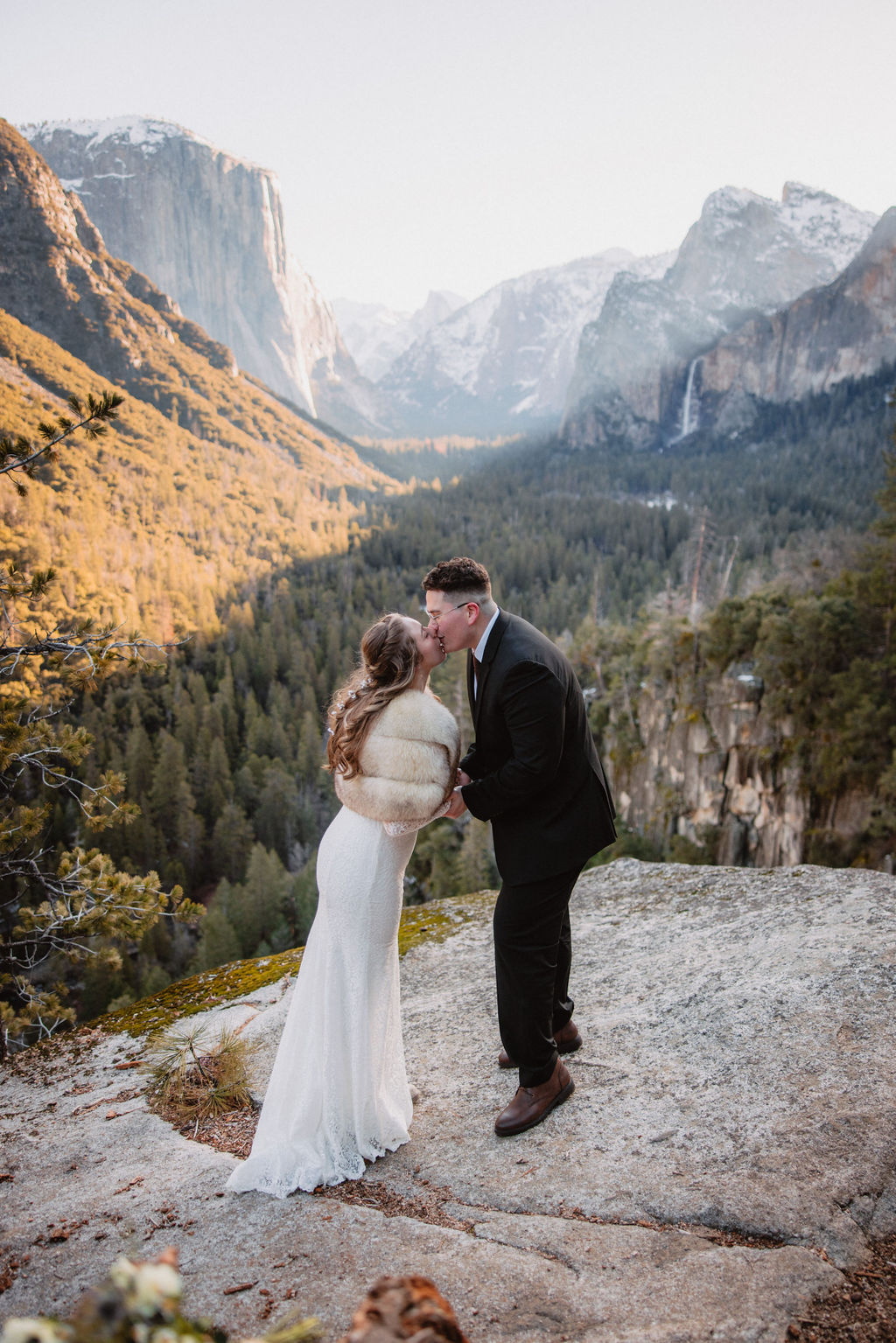 A couple embraces on a rocky overlook with a forest and mountain landscape in the background for their yosemite winter wedding