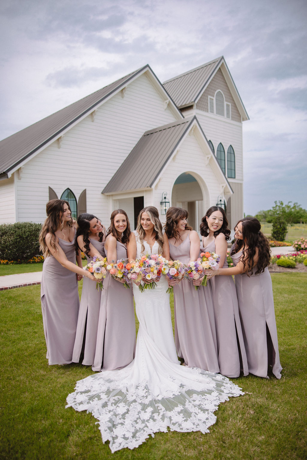 A bride in a white dress poses with six bridesmaids in gray dresses, standing together on a lawn in front of a large house.