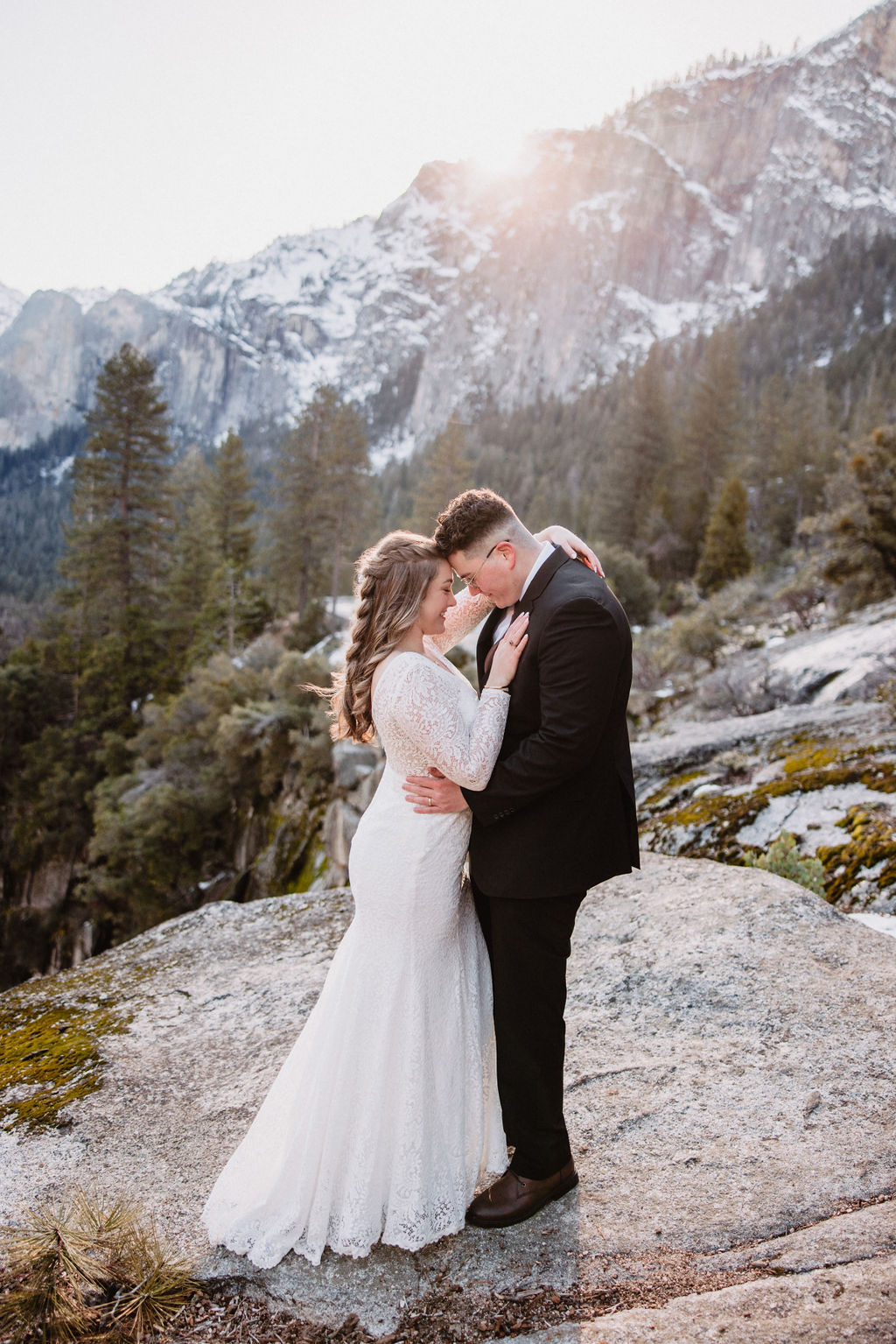 A couple embraces on a rocky overlook with a forest and mountain landscape in the background for their yosemite winter wedding