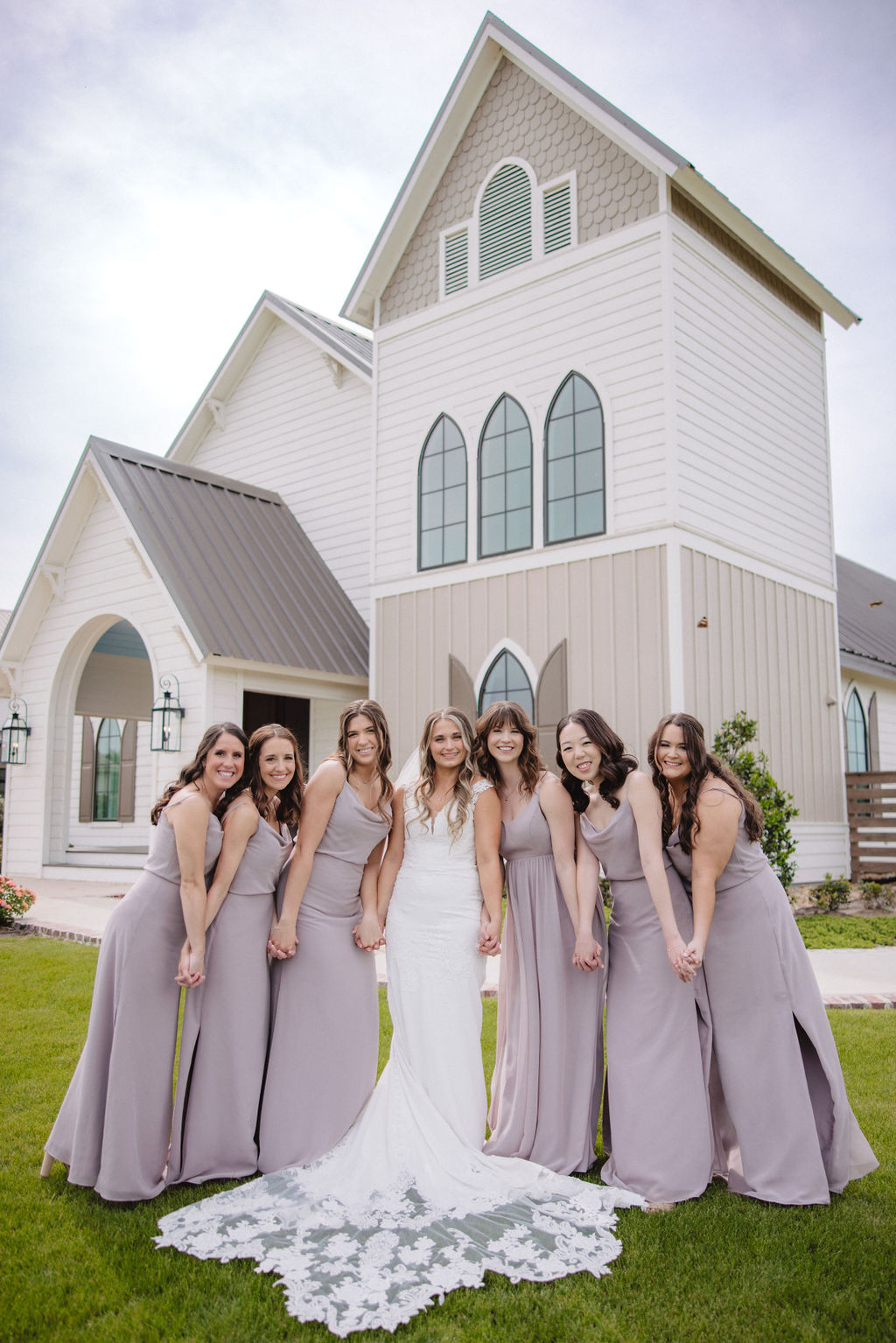 A bride in a white dress poses with six bridesmaids in gray dresses, standing together on a lawn in front of a large house.