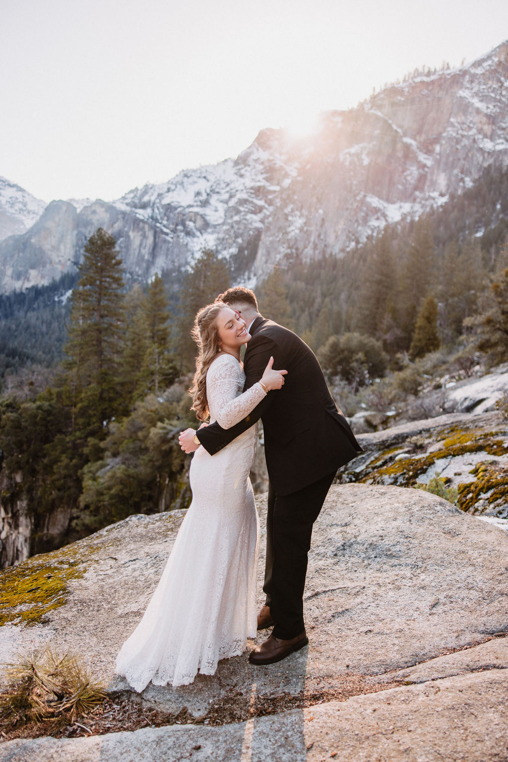 A couple embraces on a rocky overlook with a forest and mountain landscape in the background for their yosemite winter wedding