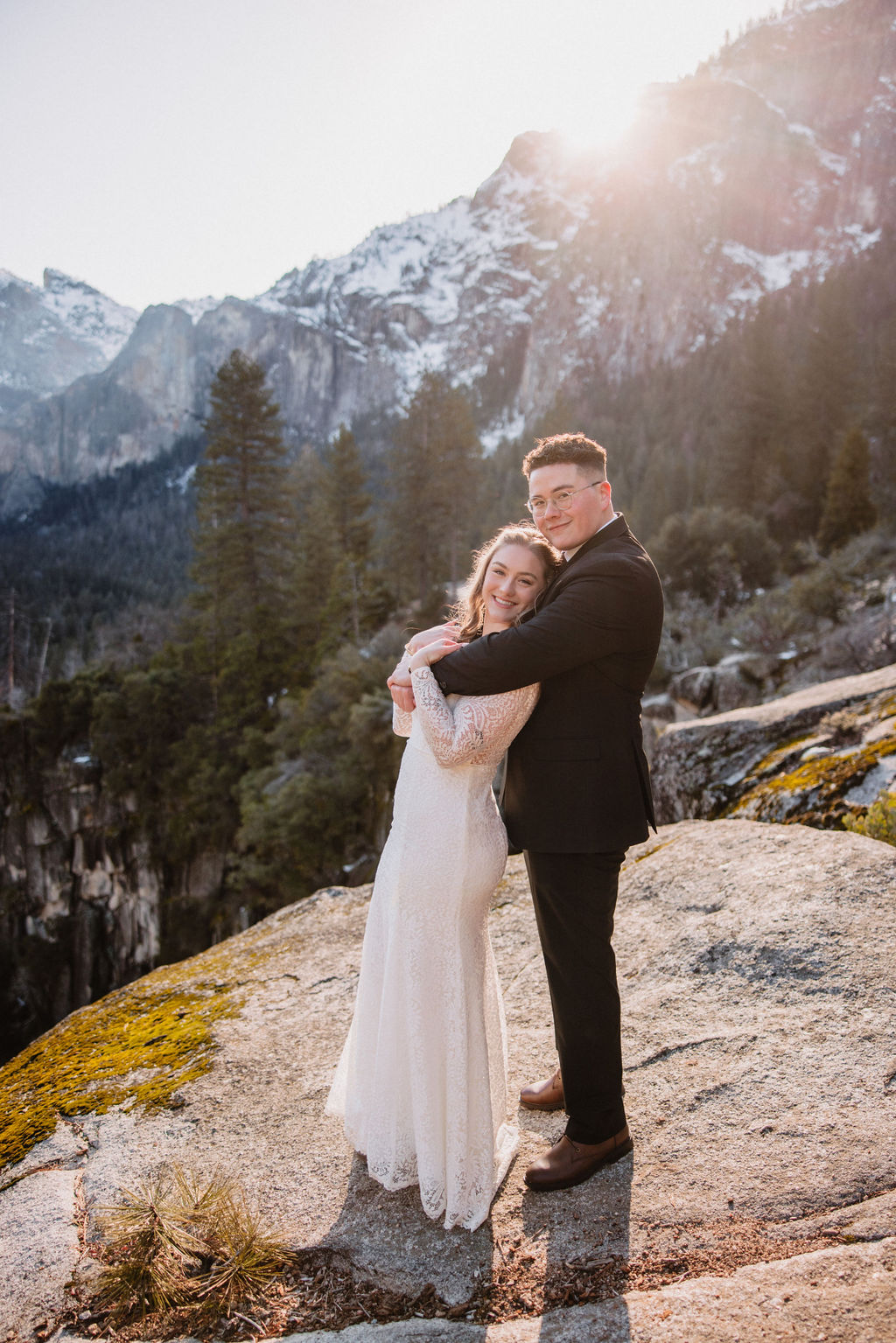 A couple embraces on a rocky overlook with a forest and mountain landscape in the background for their yosemite winter wedding