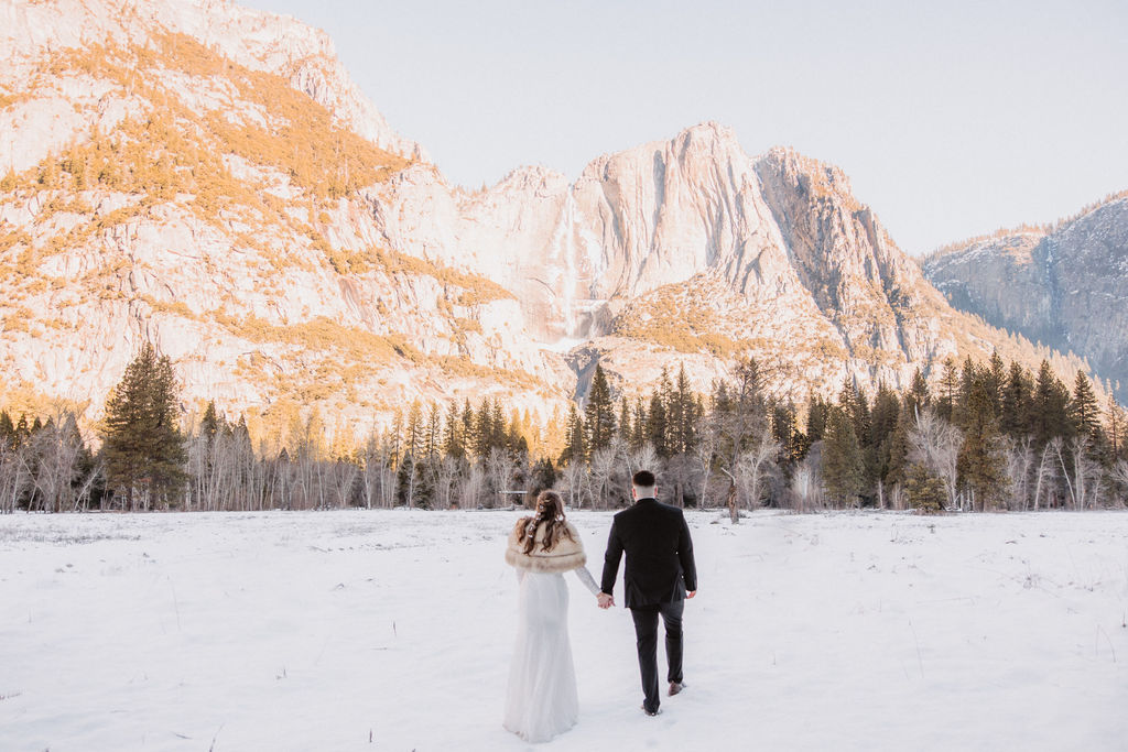 A couple in wedding attire walks hand in hand across a snowy field towards sunlit mountains under a clear sky for their yosemite winter wedding