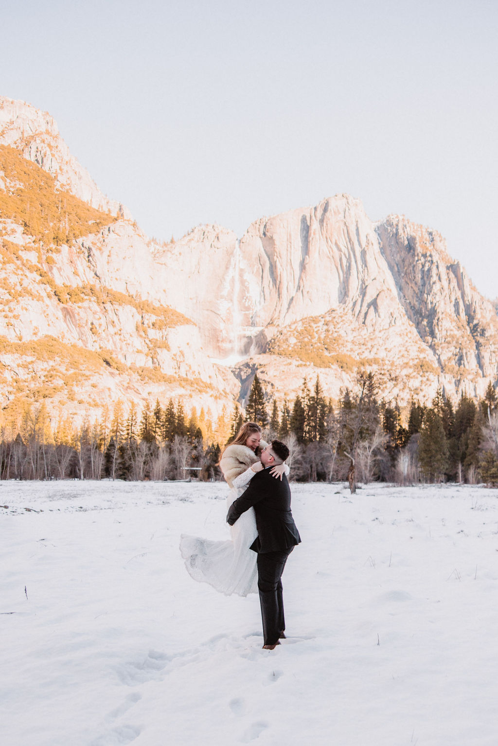 A couple in wedding attire walks hand in hand across a snowy field towards sunlit mountains under a clear sky for their yosemite winter wedding