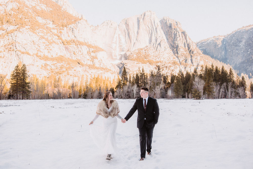 A couple is standing by a large tree; the woman is in a white dress with a fur shawl, and the man is in a dark suit. Snow is on the ground.