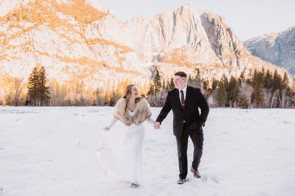 A couple is standing by a large tree; the woman is in a white dress with a fur shawl, and the man is in a dark suit. Snow is on the ground.