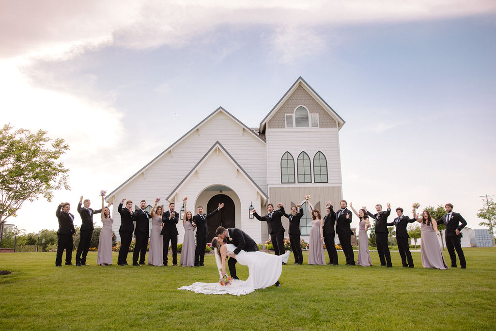 A couple kisses in front of a church, surrounded by a cheering wedding party dressed in black and gray. The Difference Between an Elopement and a Traditional Wedding