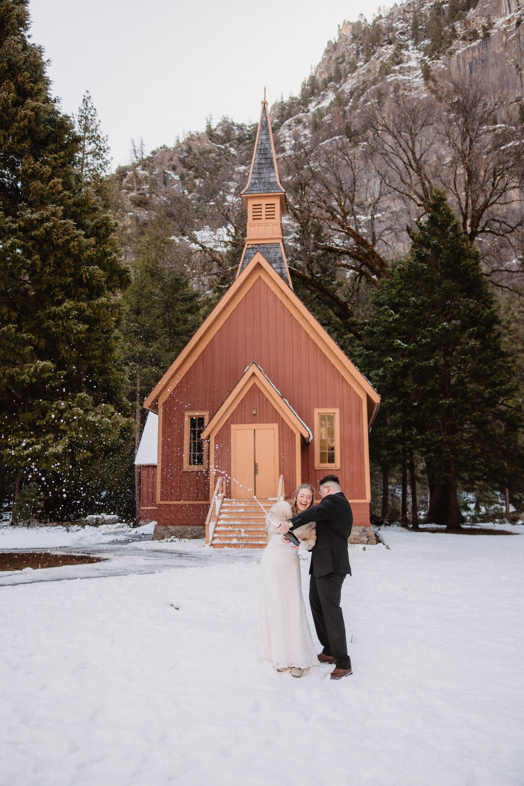 A couple stands in front of a small, brown wooden chapel surrounded by snow and trees.