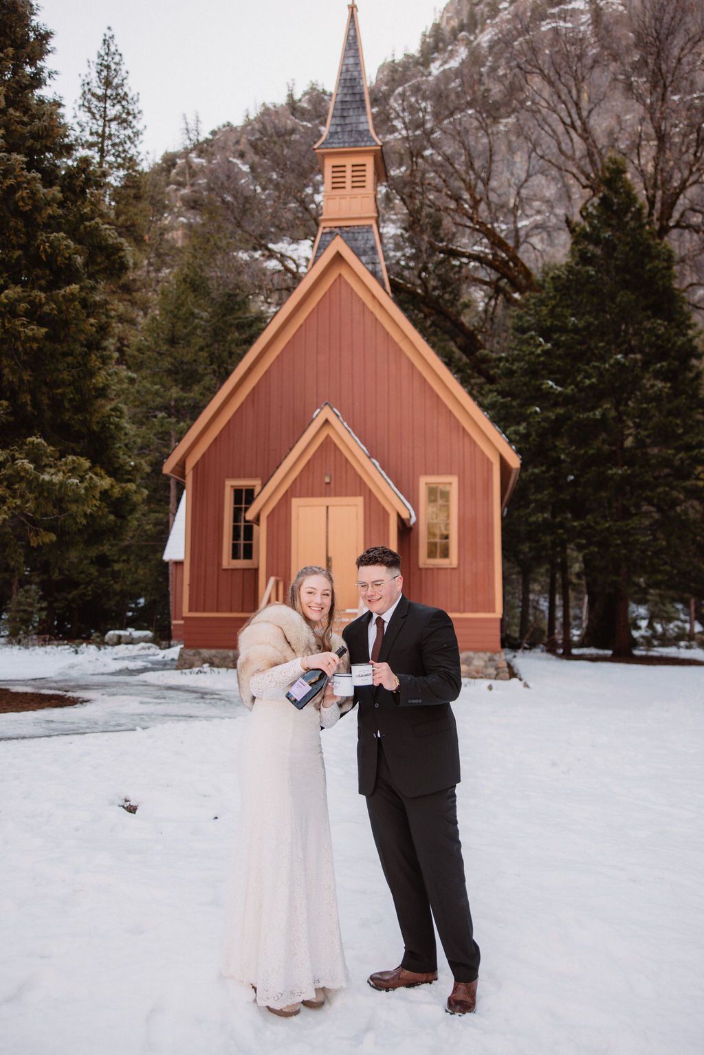 A couple stands in front of a small, brown wooden chapel surrounded by snow and trees.