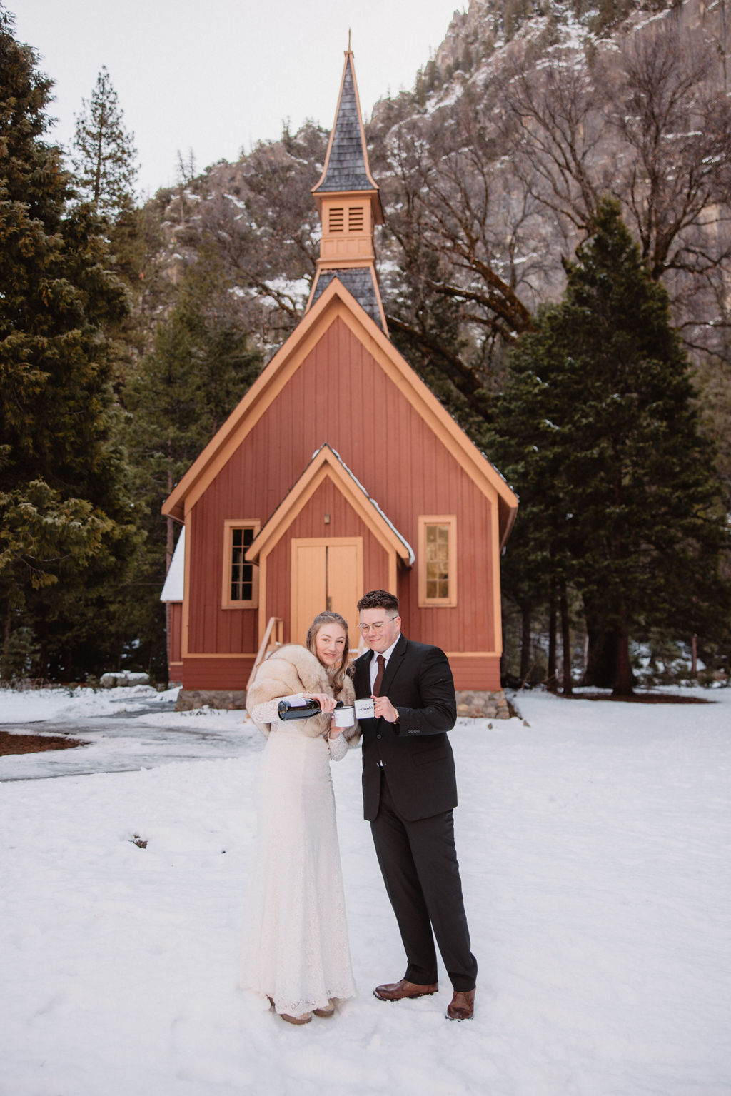 A couple stands in front of a small, brown wooden chapel surrounded by snow and trees.