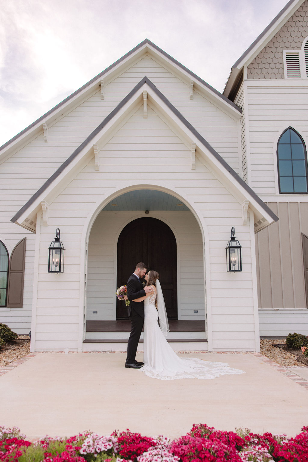 A bride and groom stand together outside a white chapel with arched doors and windows, holding hands.