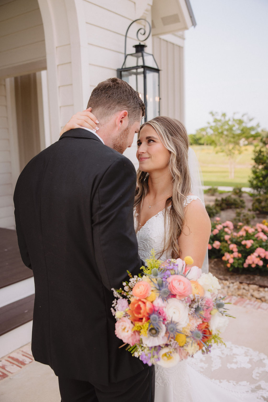 A bride and groom stand together outside a white chapel with arched doors and windows, holding hands.