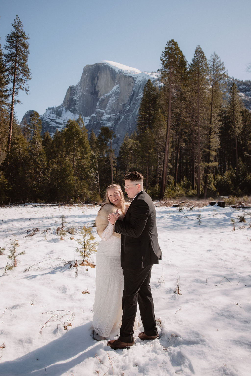 A couple in wedding attire walks hand in hand across a snowy field towards sunlit mountains under a clear sky 