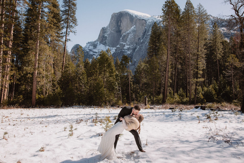 A couple in wedding attire walks hand in hand across a snowy field towards sunlit mountains under a clear sky for their yosemite winter wedding