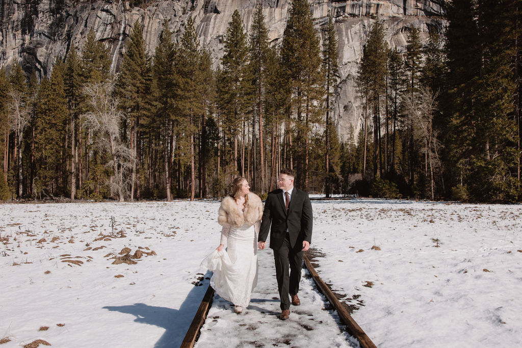 A couple in wedding attire walks hand in hand across a snowy field towards sunlit mountains under a clear sky 