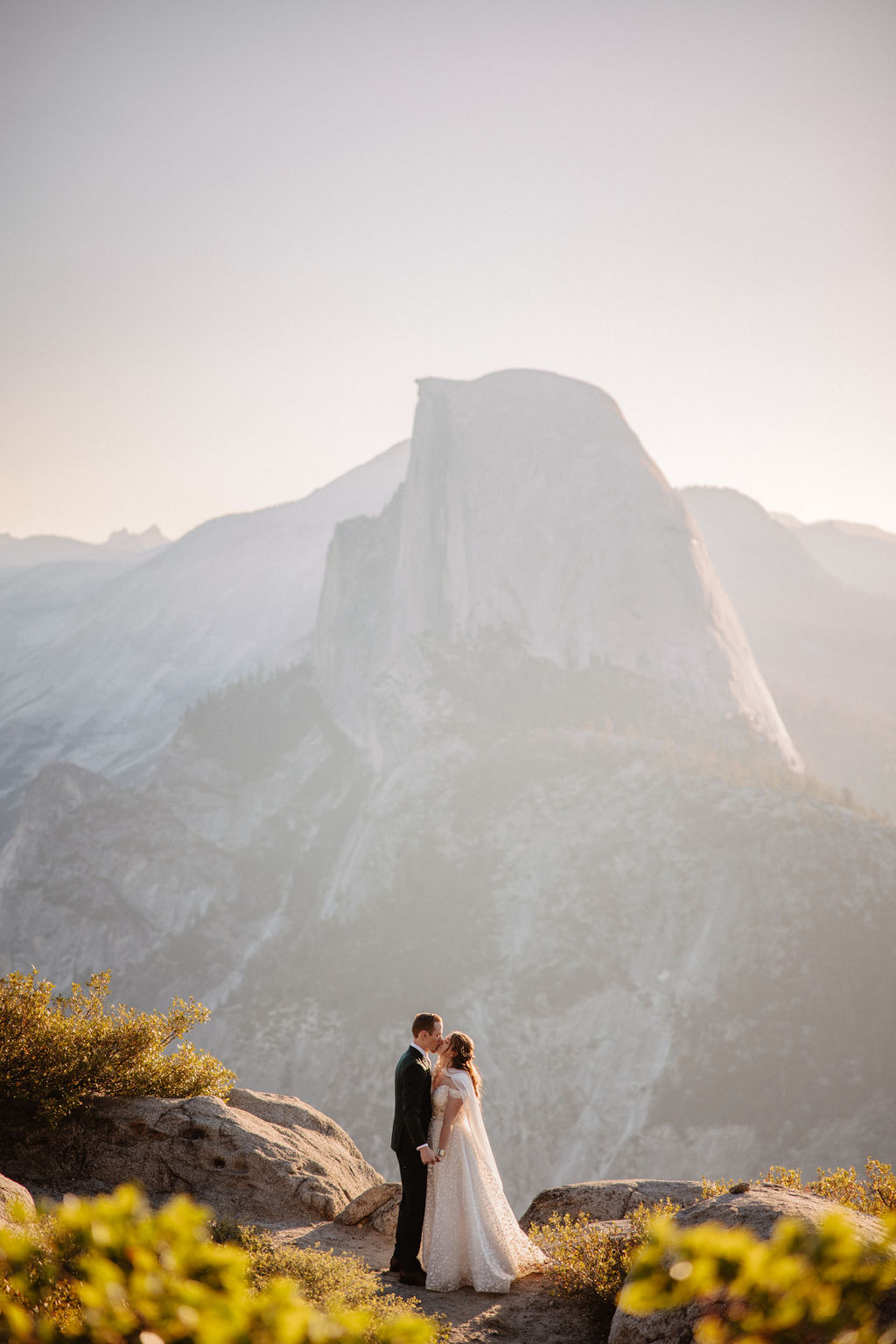 A bride and groom stand on a rocky terrain with a large mountain in the background under a clear sky.