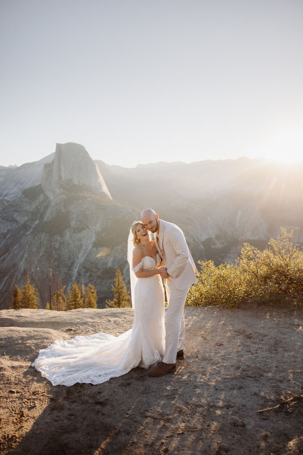 A bride and groom stand on a rocky terrain with a large mountain in the background under a clear sky.