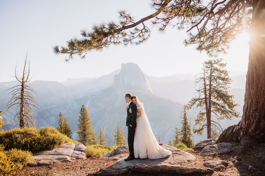 A bride and groom stand on a rocky terrain with a large mountain in the background under a clear sky.