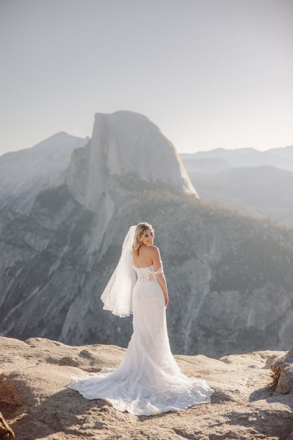 Bride in a white gown and veil stands on a rocky outcrop, with a mountain landscape in the background. |What to Wear for Your Yosemite Elopement