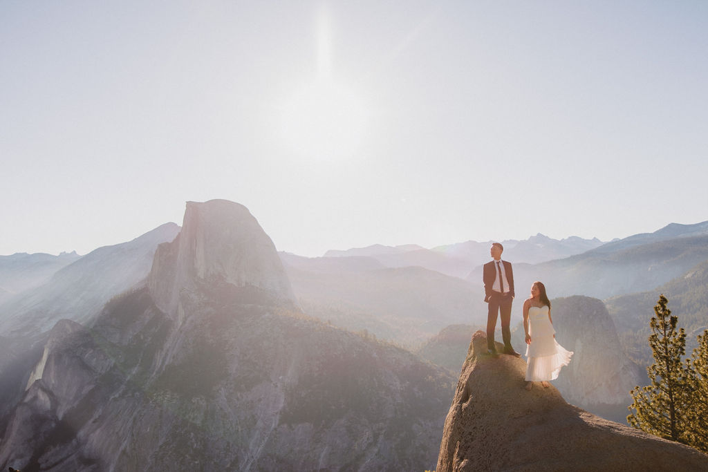 A couple stands on a cliff edge with mountain views in the background under a bright sun.