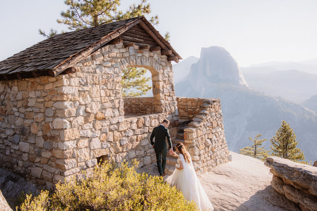 A couple in wedding attire walk towards a stone building with mountains in the background for sustainable weddings