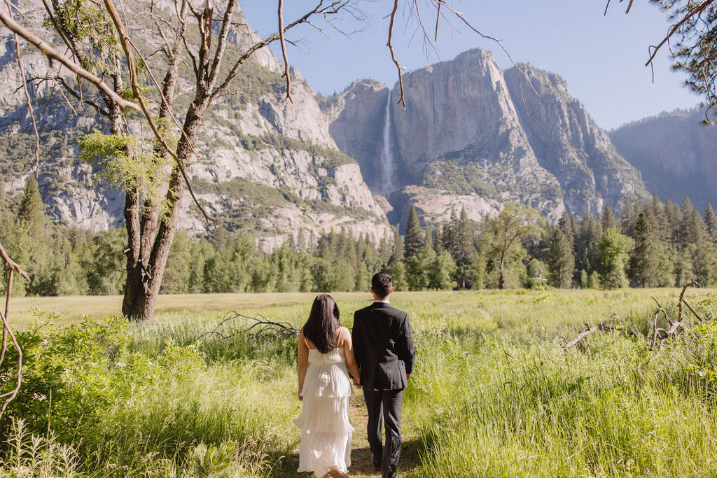 Bride and groom walking on a forest path beside a river with tall trees and mountains in the background under a clear sky.