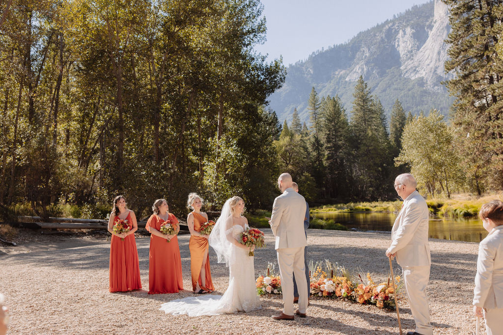 Wedding ceremony outdoors with a mountain and trees in the background. The couple stands with the officiant and two attendants, while guests are seated watching: sustainable wedding tips