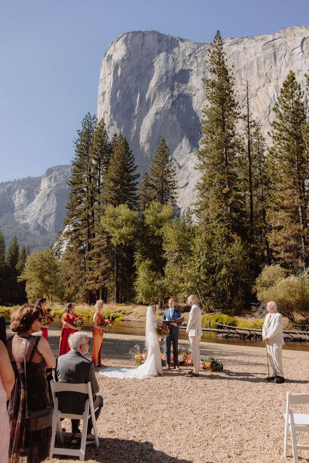 Wedding ceremony outdoors with a mountain and trees in the background. The couple stands with the officiant and two attendants, while guests are seated watching: sustainable wedding tips