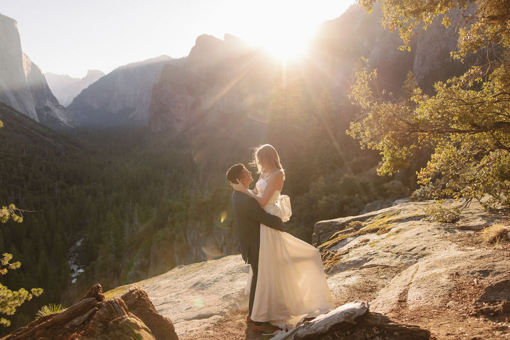 A couple embraces on a sunlit cliff with a scenic view of mountains and trees in the background.