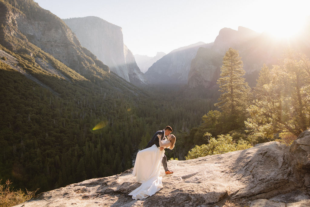 A couple embraces on a sunlit cliff with a scenic view of mountains and trees in the background.