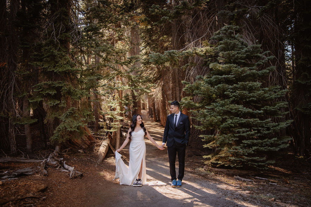 A couple embraces in a forested area, with tall trees surrounding them and sunlight filtering through the branches. | What to Wear for Your Yosemite Elopement