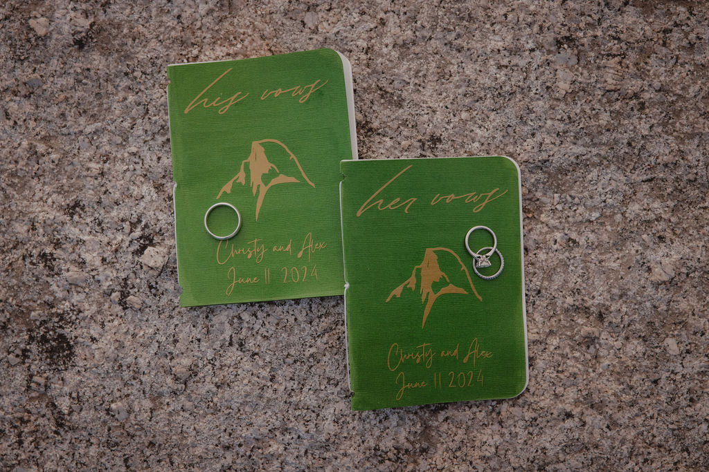 Two green notebooks with illustrations of a mountain on a stone surface. Each notebook has a metal ring placed on it.