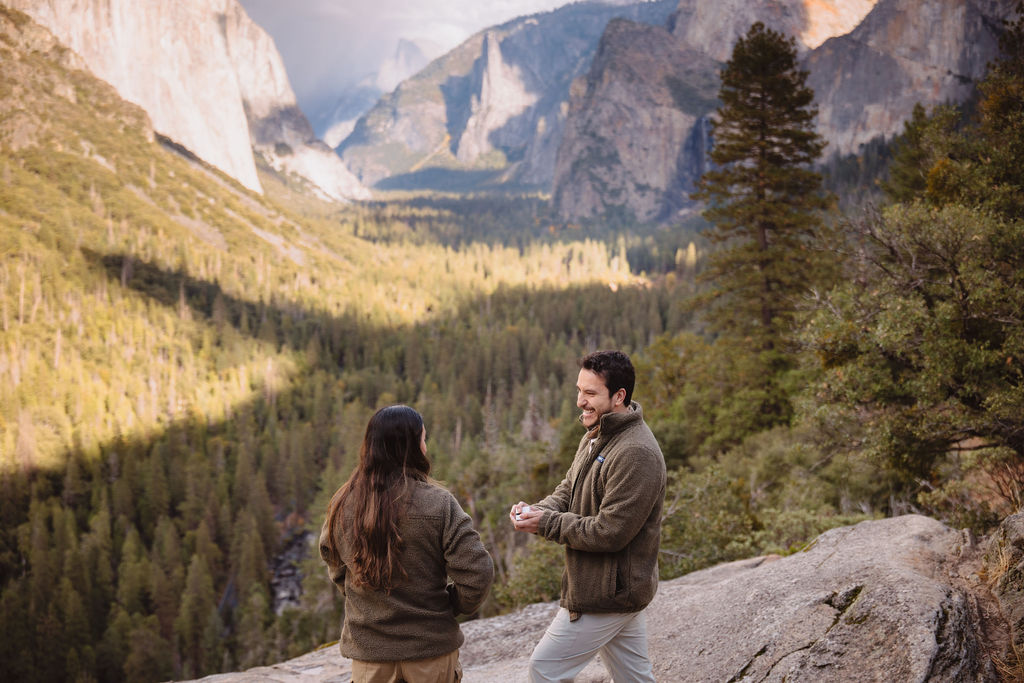 A person kneels and holds hands with another, set against a mountainous landscape with a valley and cloudy sky taken by a yosemite proposal photo