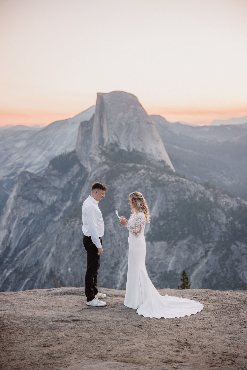 A couple in wedding attire stands on a cliff ledge with a large rock formation in the background, under a pastel sky.