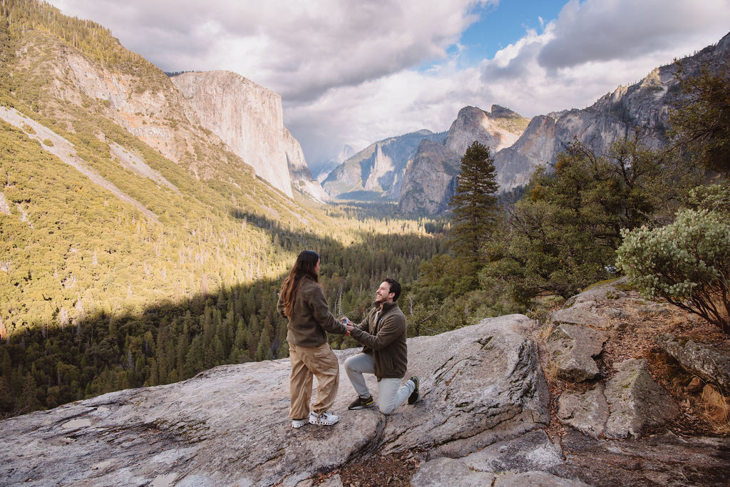 A person kneels and holds hands with another, set against a mountainous landscape with a valley and cloudy sky taken by a yosemite proposal photo