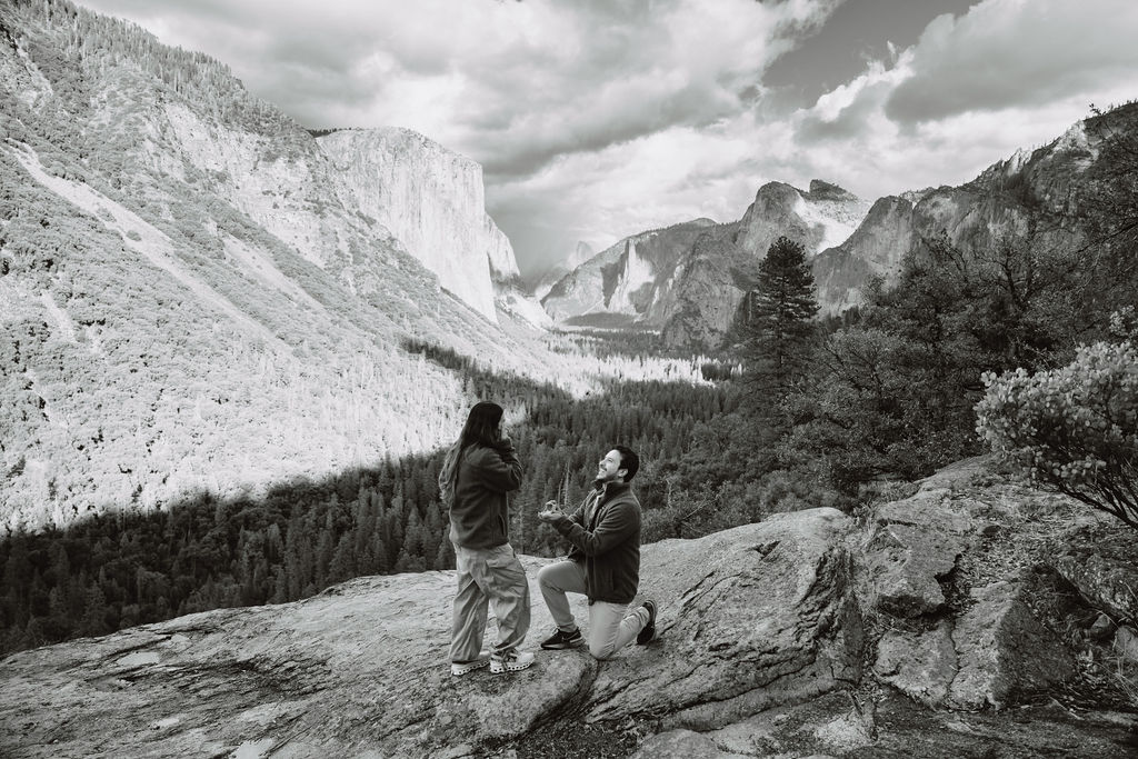 A person kneels and holds hands with another, set against a mountainous landscape with a valley and cloudy sky taken by a yosemite proposal photo