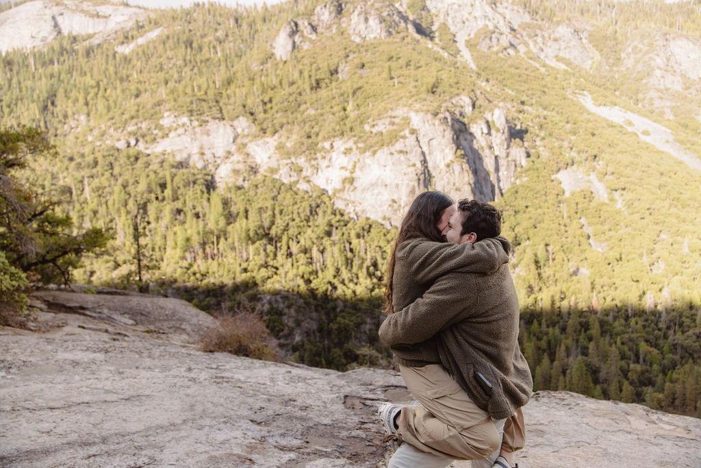 A person kneels and holds hands with another, set against a mountainous landscape with a valley and cloudy sky taken by a yosemite proposal photo