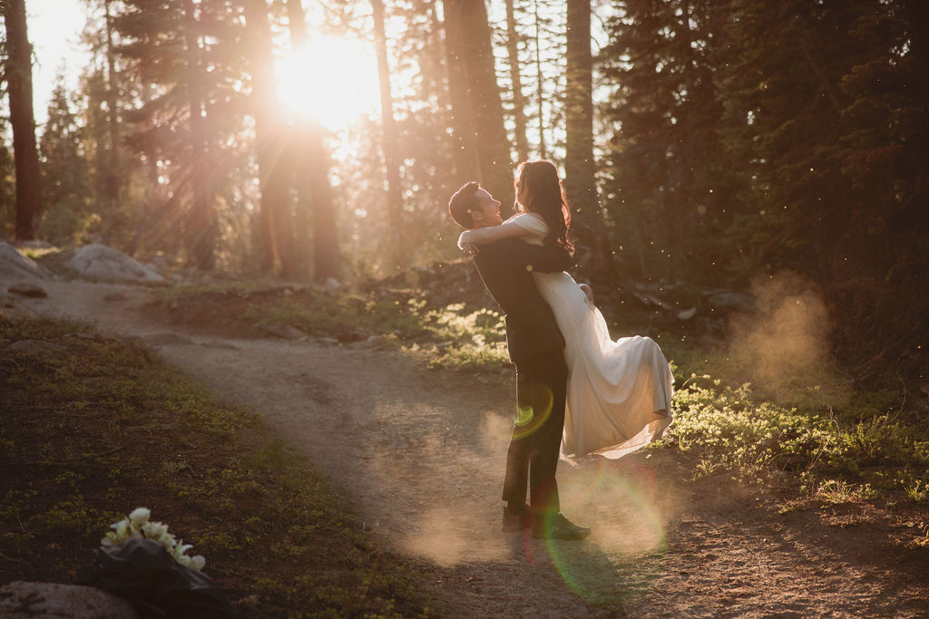 A couple walks hand in hand on a forest path during sunset. The woman wears a flowing dress, and the man is in a suit. Sunlight filters through the trees: sustainable wedding tips