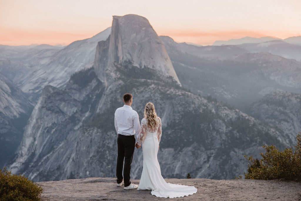 A couple in wedding attire stands on a cliff ledge with a large rock formation in the background, under a pastel sky.